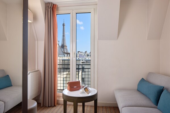 A hotel room with a view of the Eiffel Tower, a round table with a handbag and a photo frame, and two sofas with cushions.
