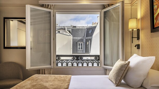 Une chambre d’hôtel avec un lit, une fenêtre donnant sur des immeubles, une chaise et une lampe fixée au mur.