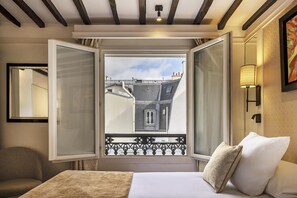 Une chambre d’hôtel avec un lit, une fenêtre donnant sur des immeubles, une chaise et une lampe fixée au mur.