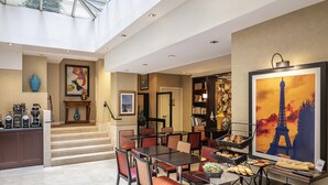 Un hall d’hôtel spacieux avec un plafond haut, un escalier menant à un étage supérieur, un coin café, un espace repas avec des tables et des chaises, et un grand tableau représentant la Tour Eiffel.