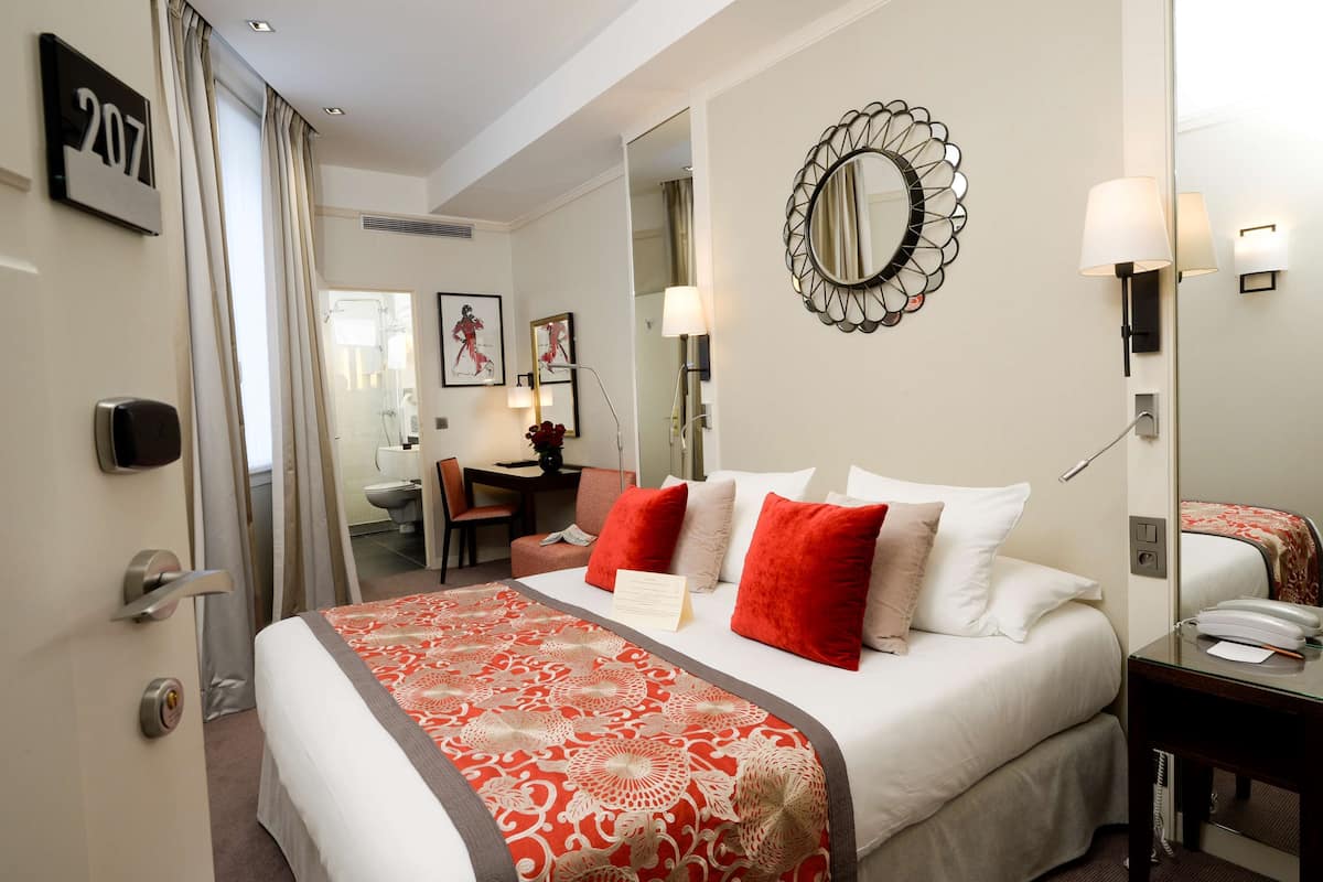 A hotel room with a bed, a desk, a chair, a bathroom, and a mirror.