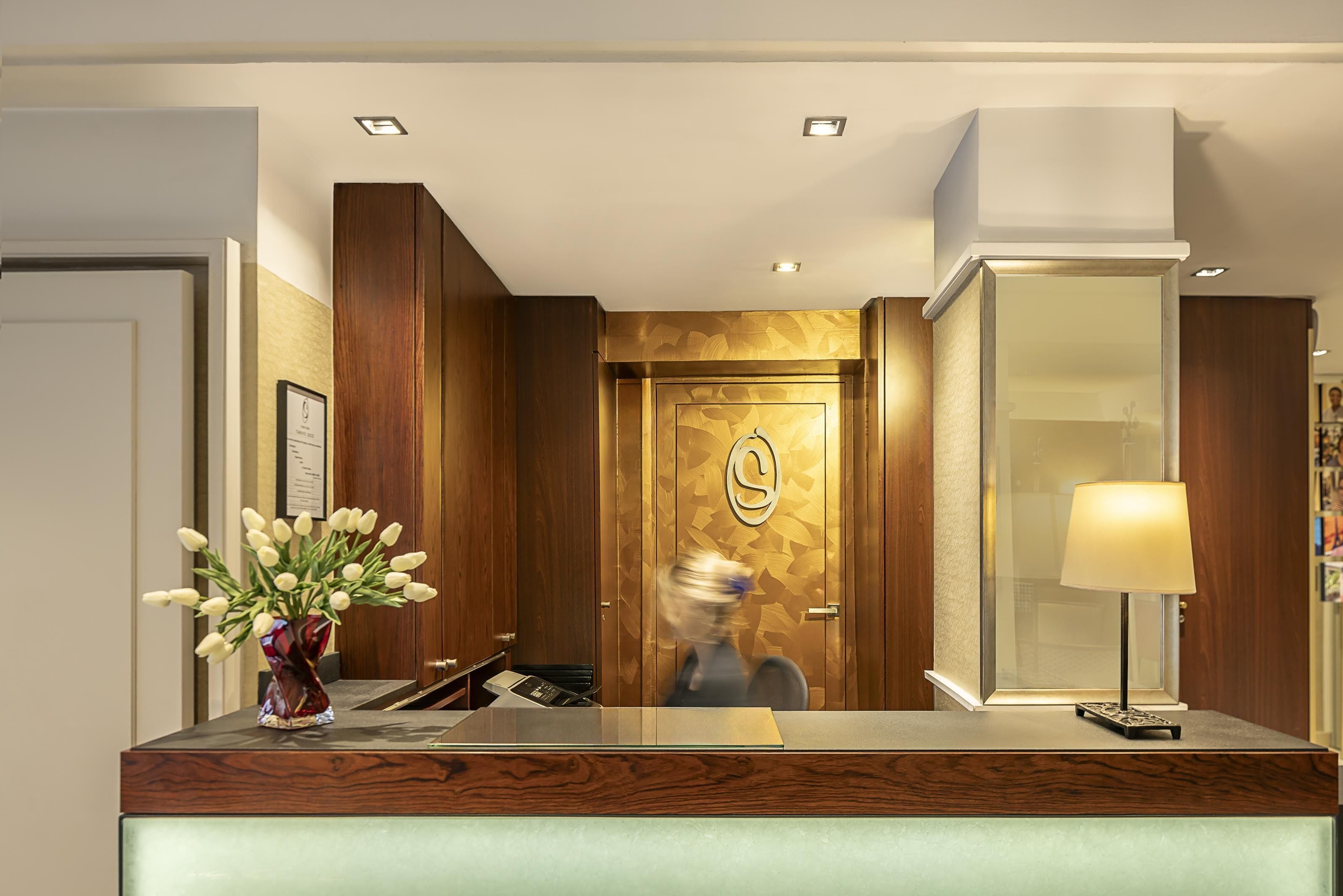 Zona de recepción de un hotel con un mostrador de madera, un jarrón de flores, una lámpara y una puerta con diseño geométrico.