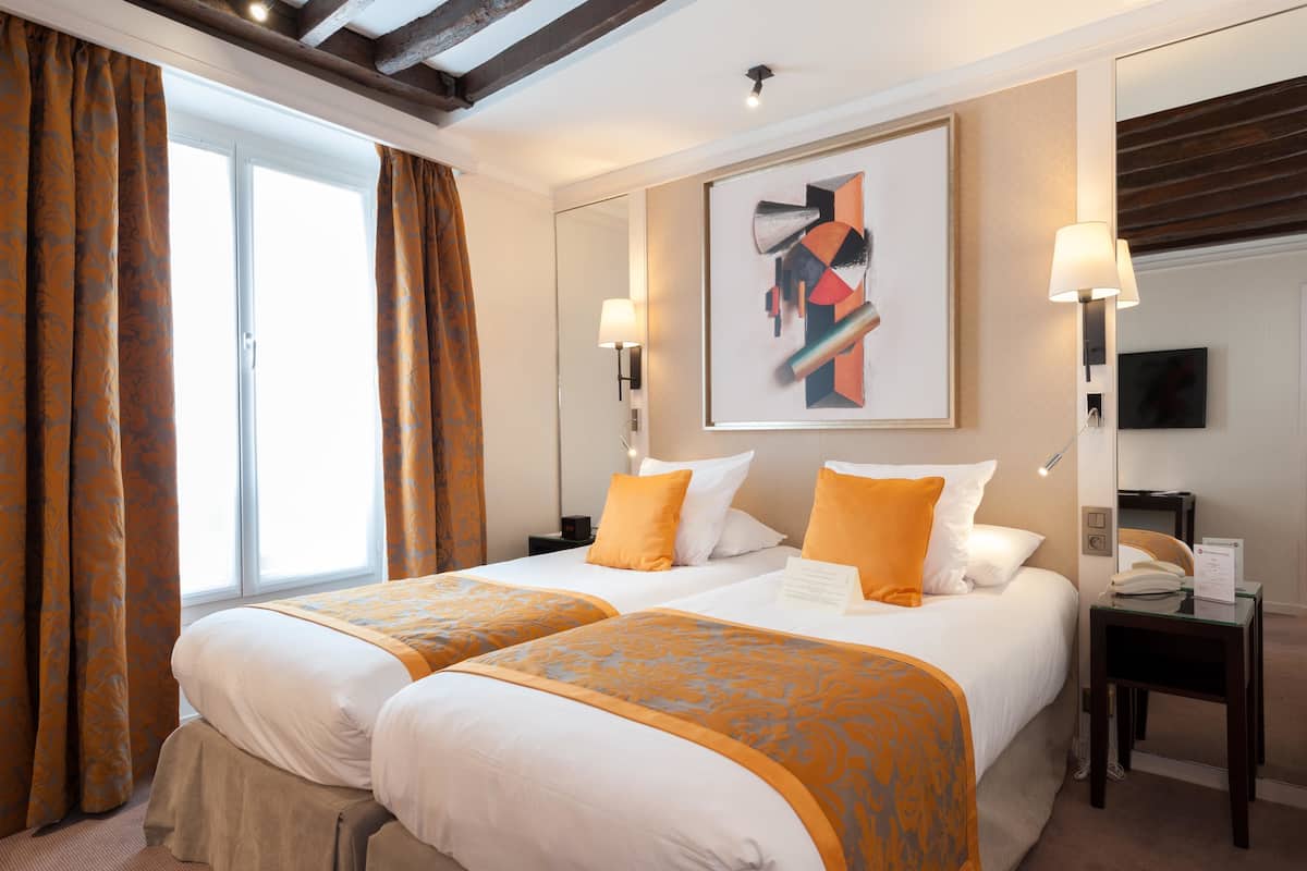A hotel room with two beds, a large window with curtains, a painting on the wall, and a desk with a lamp.