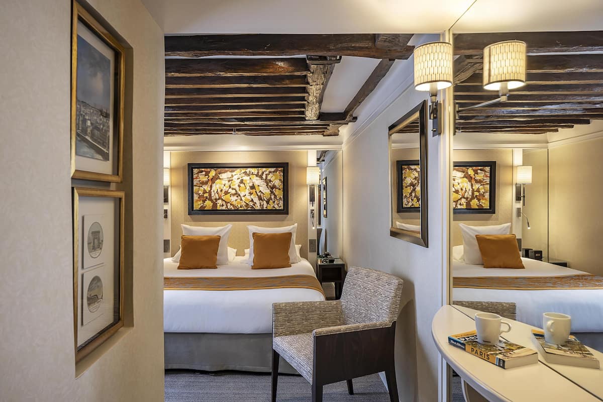 A hotel room with a large bed, a chair, a desk with a mirror, and artwork on the walls.
