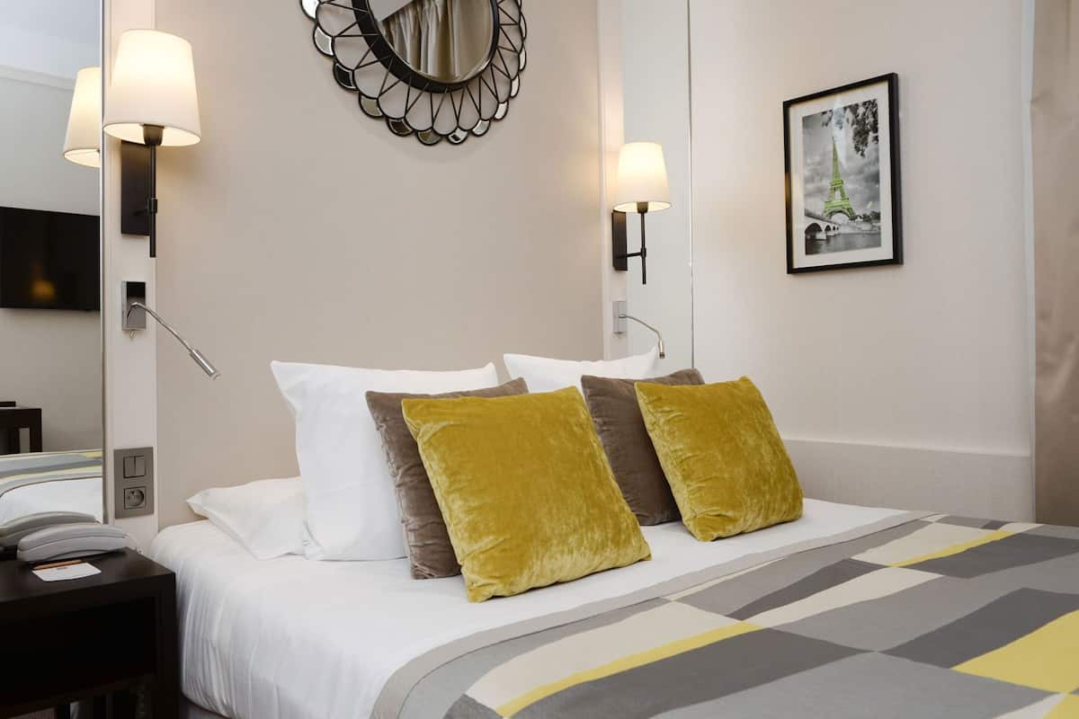 A hotel room with a bed, two wall-mounted lamps, a framed picture of the Eiffel Tower, and a bedside telephone.