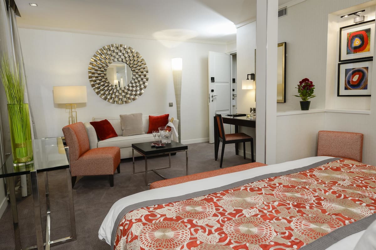 A hotel room with a bed, a sofa, a desk, and a chair. There is a mirror on the wall and a vase with flowers.