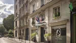 La façade d’un hôtel, ornée d’une enseigne indiquant « Sydney Opera », présente un style architectural classique avec de nombreuses fenêtres et balcons, des plantes en pot et un auvent métallique.