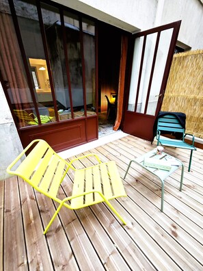 A wooden deck with yellow and green chairs, a small table, and a sliding glass door leading to a room with a bed and a nightstand. - Le Relais Monceau (Paris)