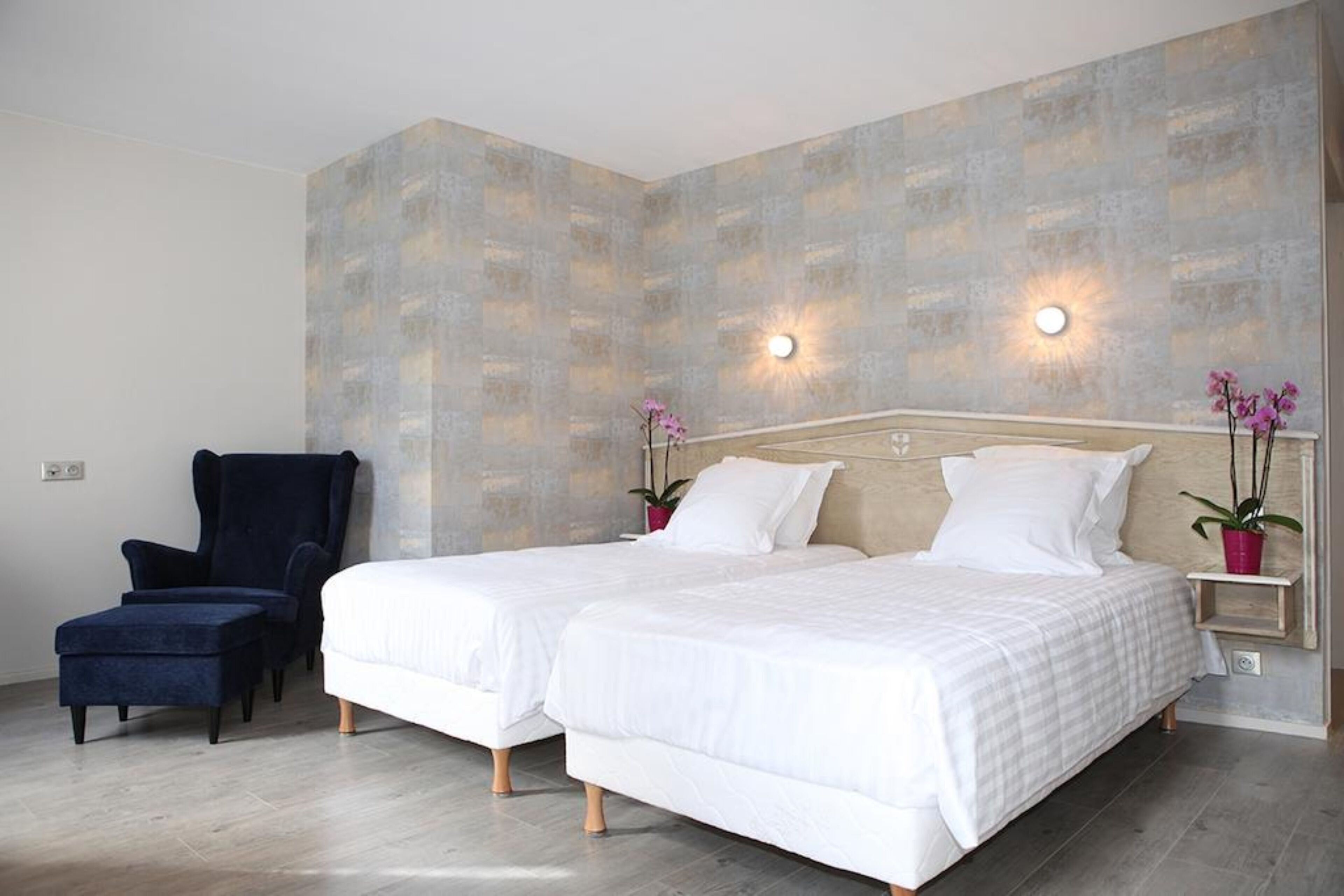 Habitación de hotel con dos camas, un sillón oscuro y una lámpara fijada a la pared.