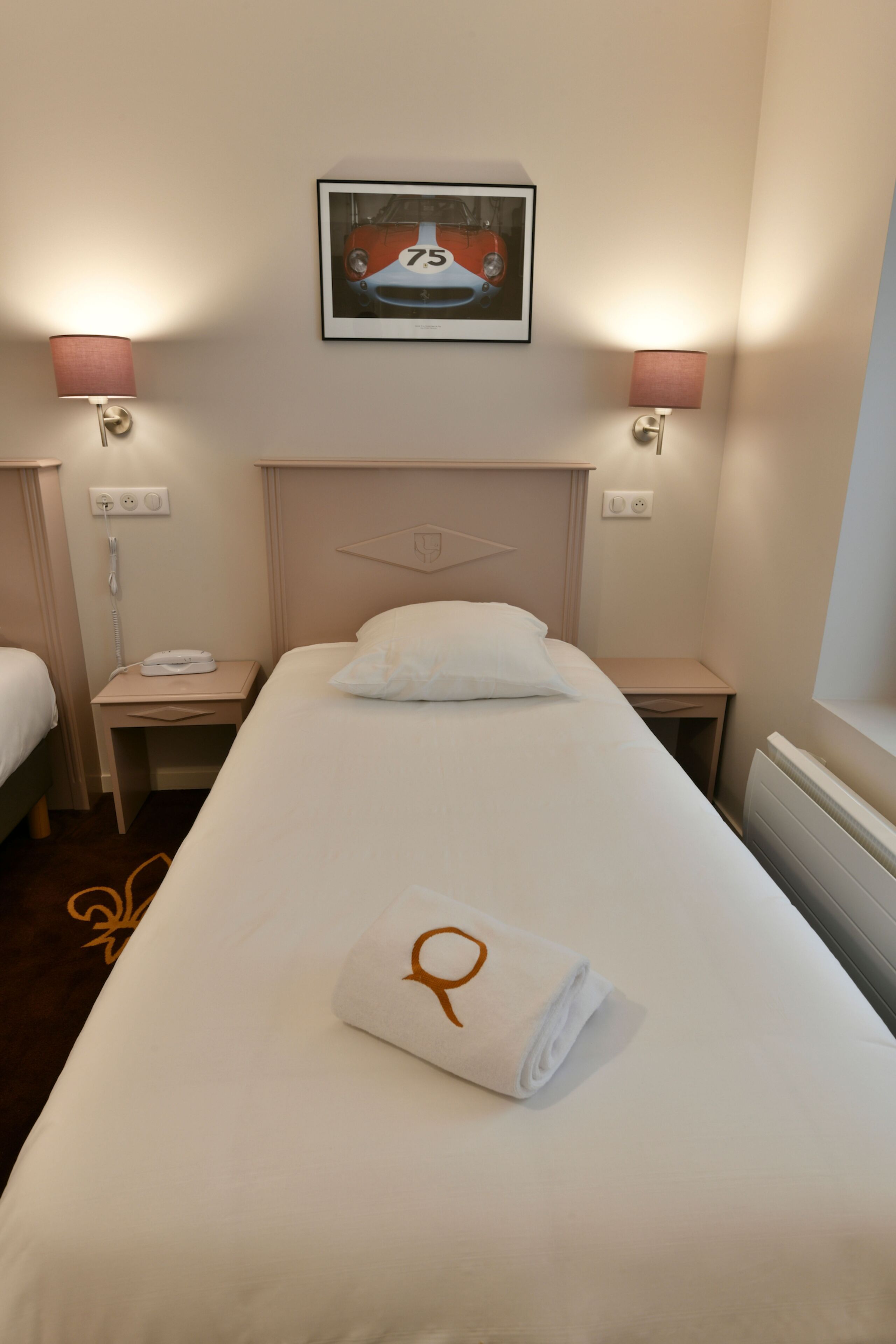 Habitación de hotel con una cama individual, una mesita de noche con lámpara, un cuadro en la pared y una toalla enrollada sobre la cama.