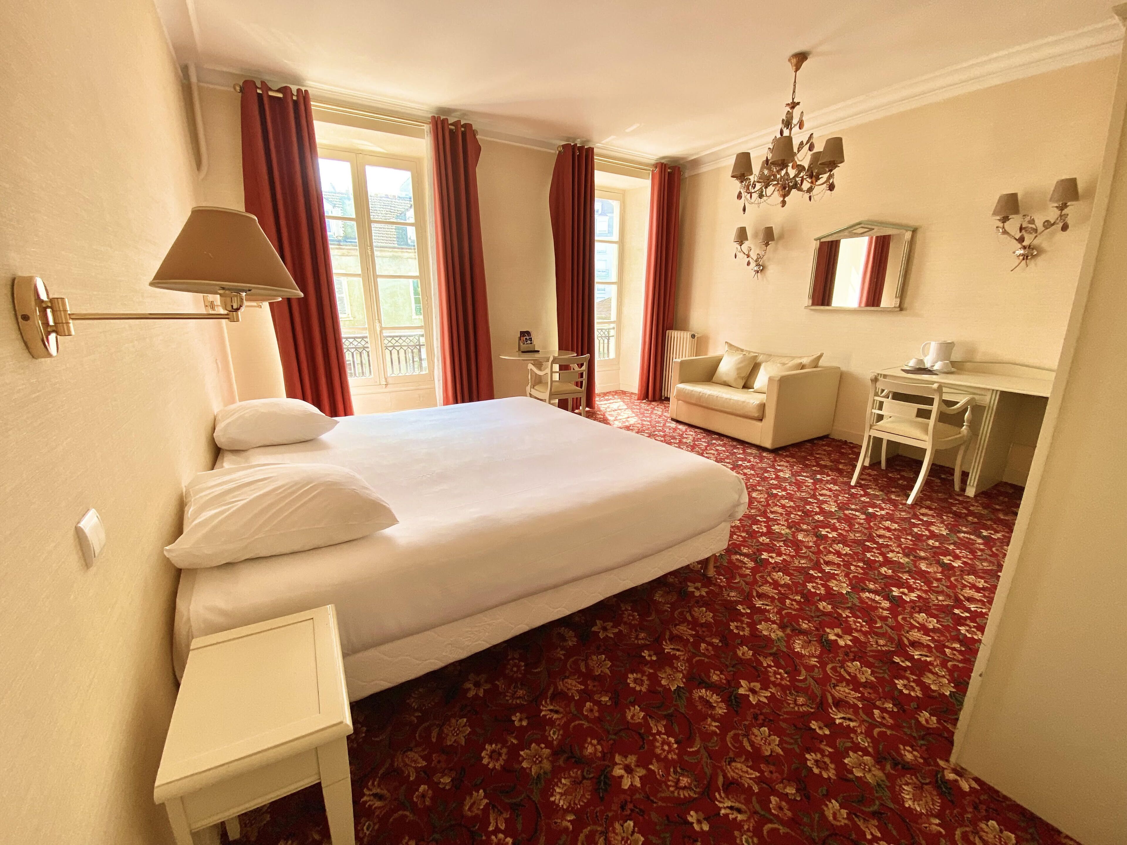 A hotel room with a large bed, a desk, a chair, a small table, a mirror, and a chandelier.