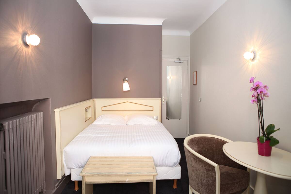 A hotel room with a bed, a chair, a small table, and a potted plant.