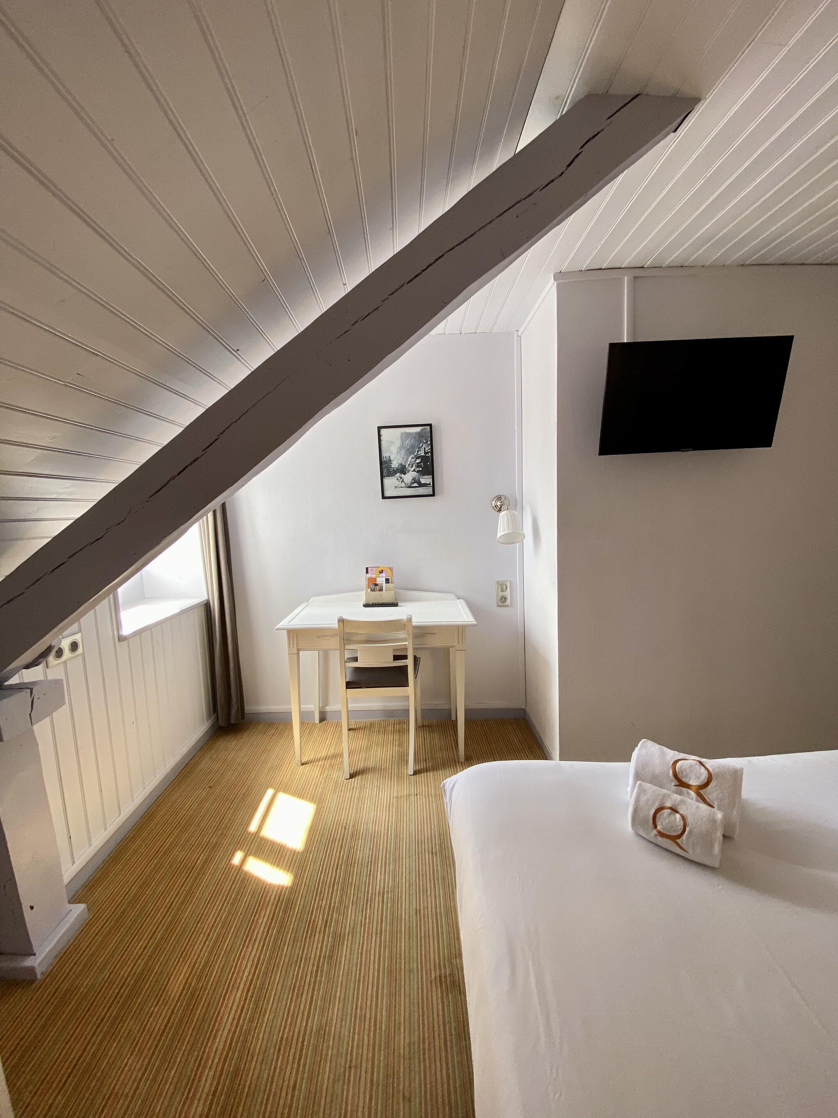 Una habitación con techo inclinado, una cama, un escritorio con silla, un televisor montado y un cuadro en la pared.