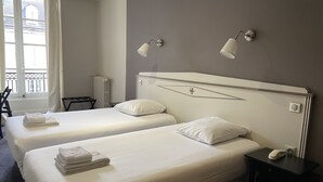 A hotel room with two beds, a bedside table, a chair, and a window with curtains.
