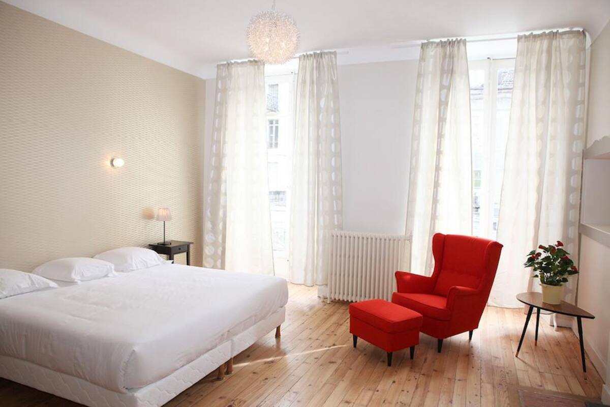A bedroom with a bed, a red chair, a small table with a plant, and a window with curtains.