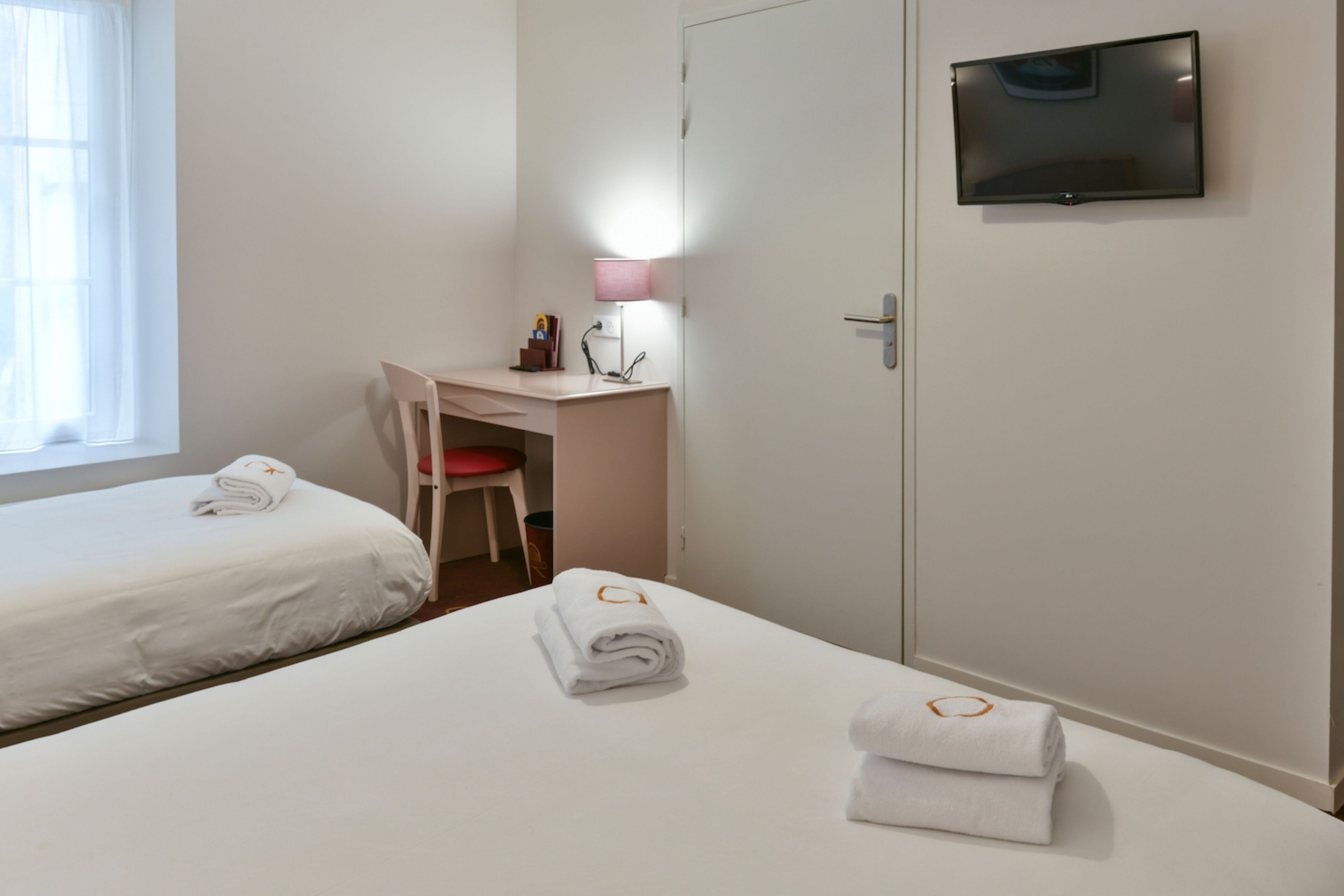 Quarto de hotel com duas camas, uma escrivaninha com cadeira, televisão na parede e uma luminária.