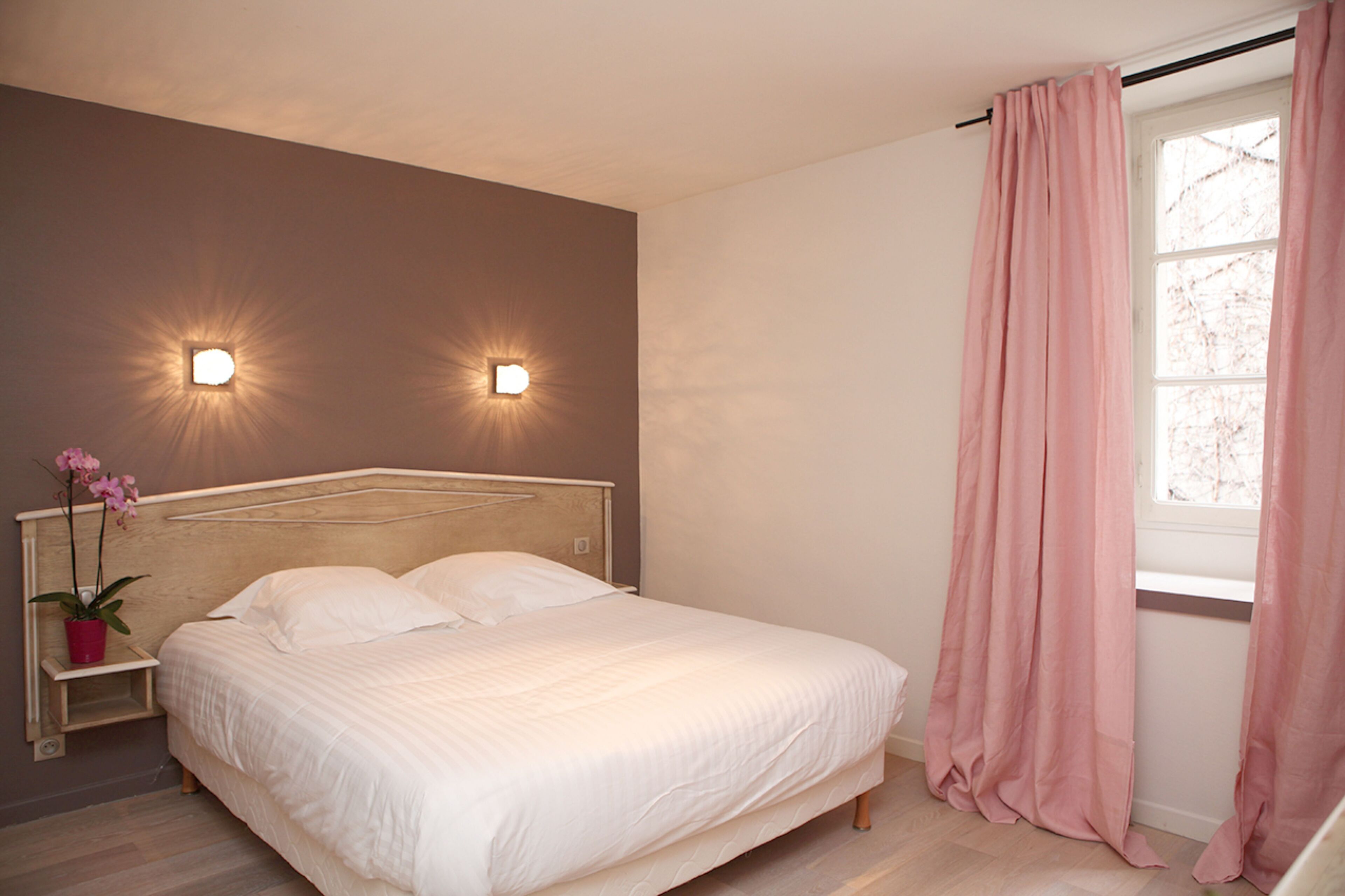 Un dormitorio con una cama, dos luces fijadas a la pared, un ventanal con cortinas rosadas y una mesita de noche con una planta.