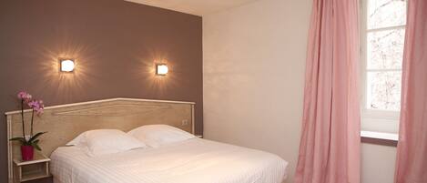 Un dormitorio con una cama, dos luces fijadas a la pared, un ventanal con cortinas rosadas y una mesita de noche con una planta.