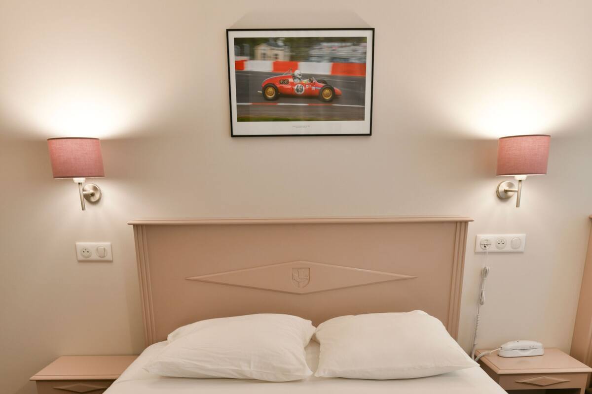 A hotel room with a single bed, two white pillows, two bedside tables with lamps, a framed picture on the wall, and two towels with a logo on them.