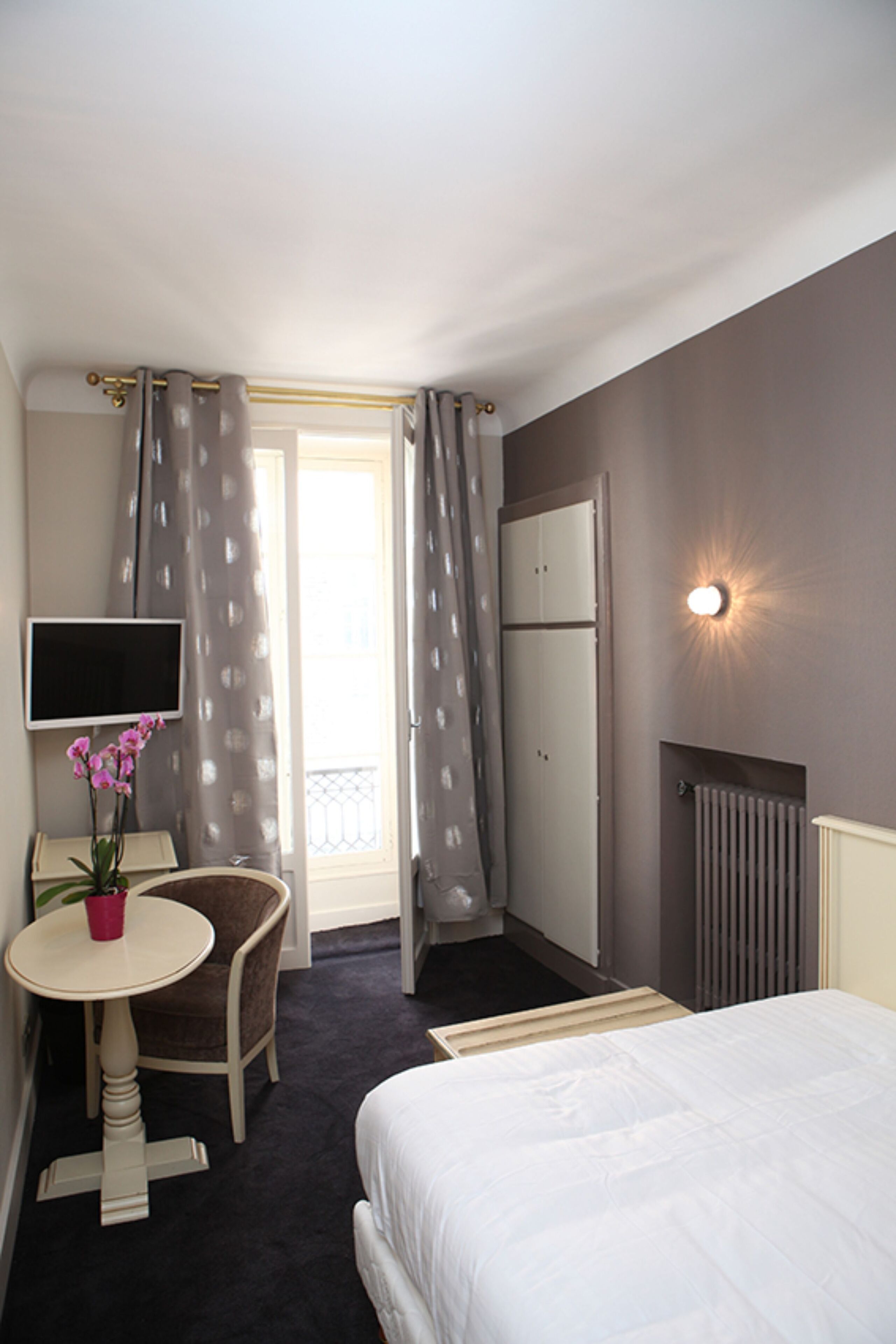 A hotel room with a bed, a desk with a chair, a TV, a potted plant, and a window with curtains.