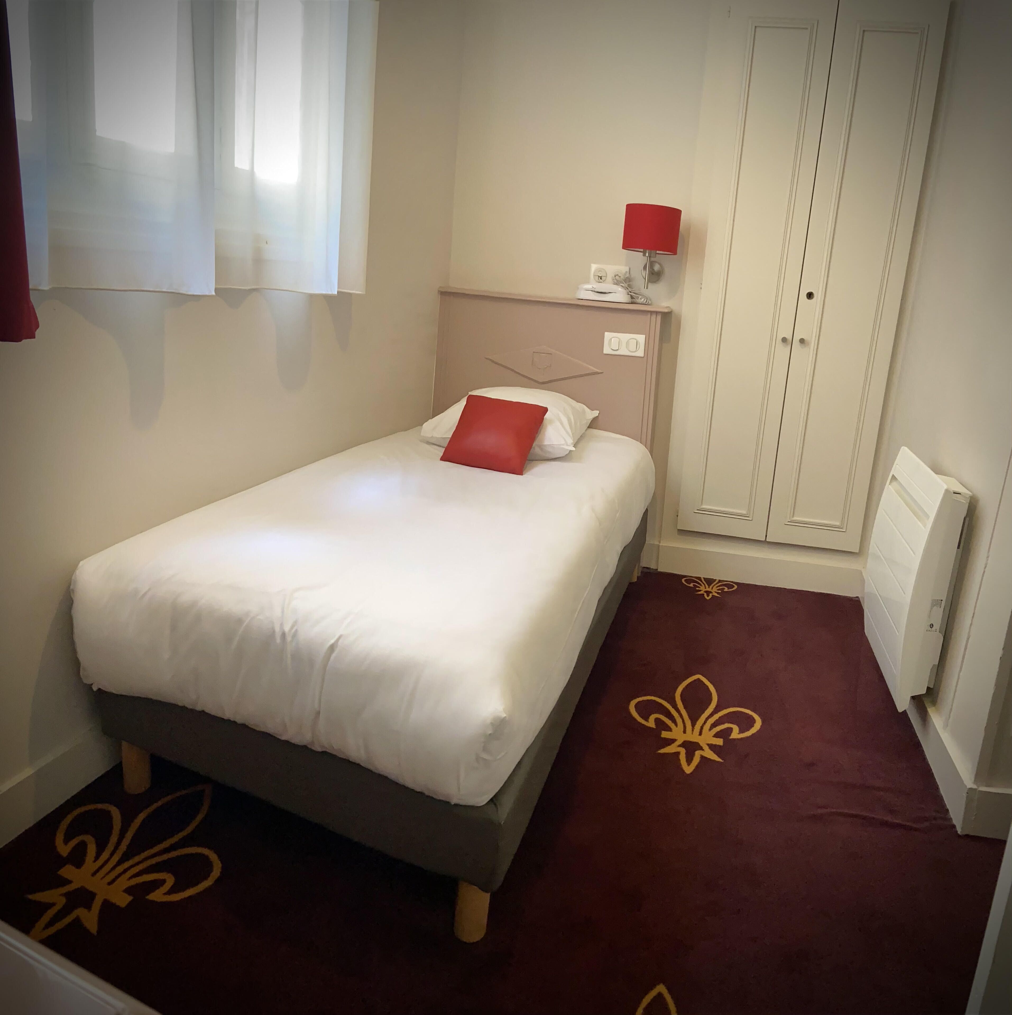 A single bed with white bedding and a red pillow, a nightstand with a lamp, a wardrobe, and a radiator on the floor.