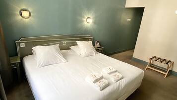 Una cama bien hecha con sábanas y toallas blancas, dos mesitas de noche con lámparas, y un pequeño perchero de madera en el suelo.