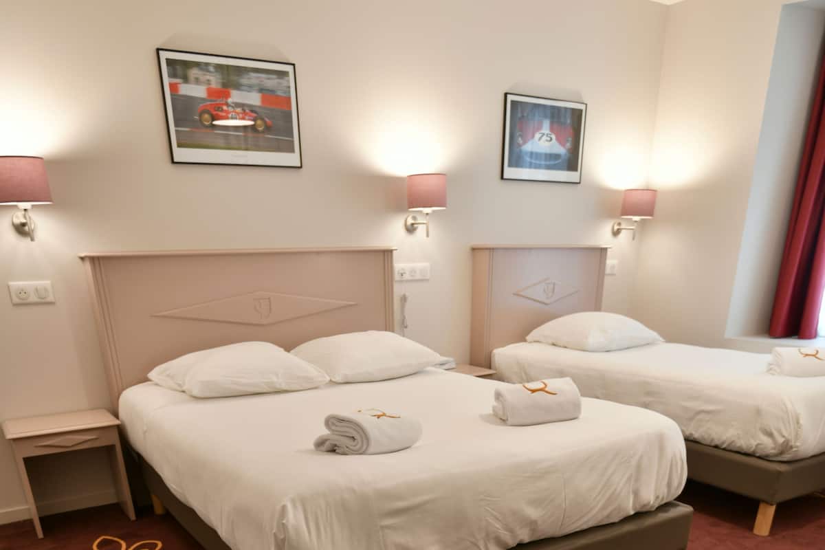 A hotel room with two beds, each with white linens and a headboard. There are wall-mounted lamps on either side of the beds, and a framed picture above each bed. The room has a brown carpet with a fleur-de-lis pattern.