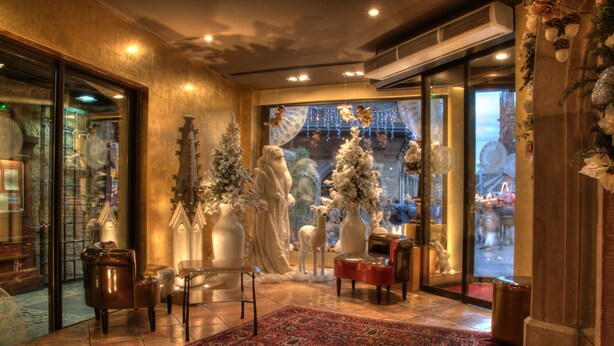 Un hall d’entrée avec un tapis à motifs rouges, une chaise en bois, une porte vitrée donnant sur l’extérieur, et des décorations de Noël, notamment une figurine du Père Noël et des arbres recouverts de neige.