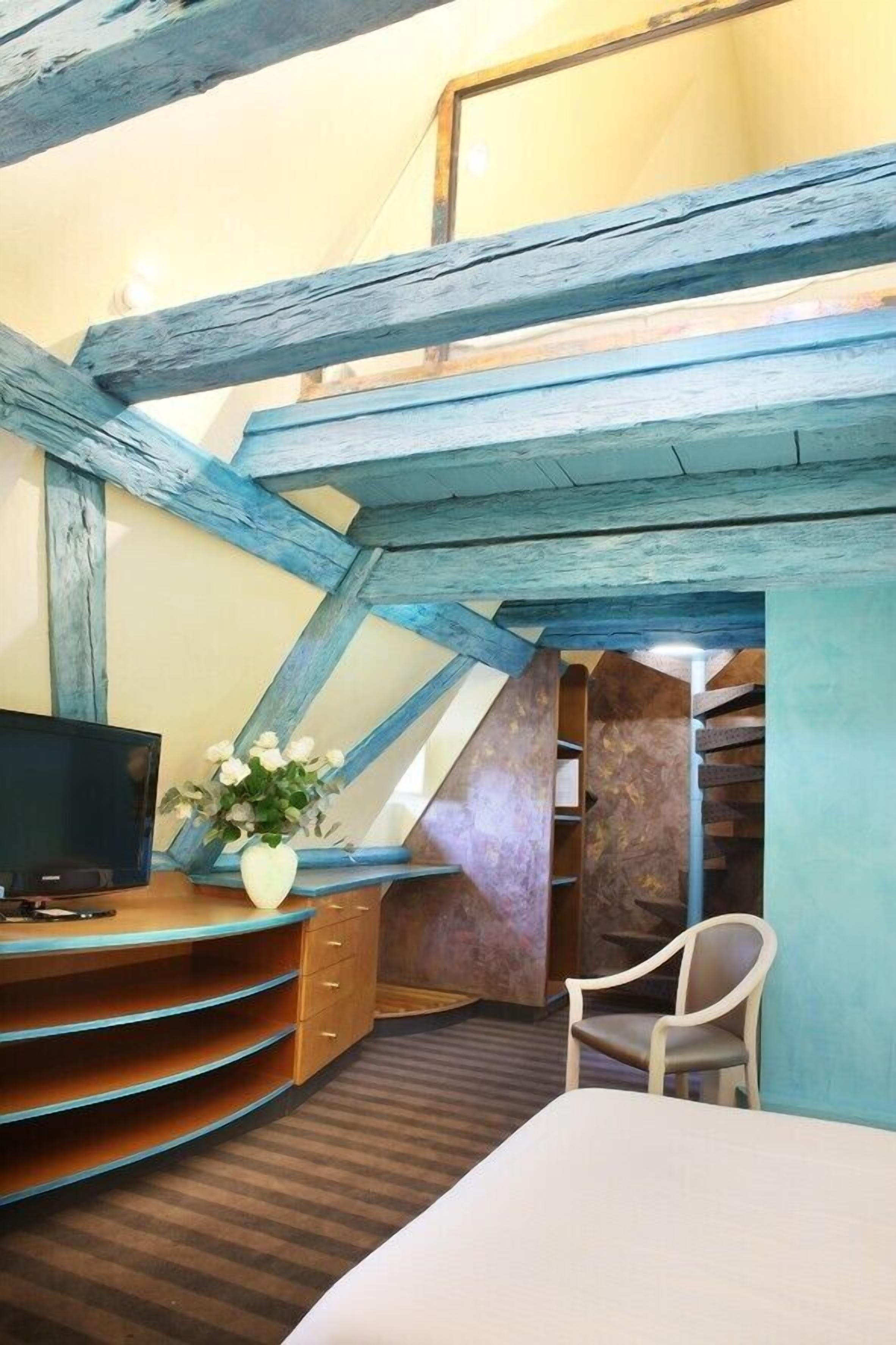 A bedroom with a sloped ceiling, a bed, a TV, a desk with a chair, and a vase with flowers.