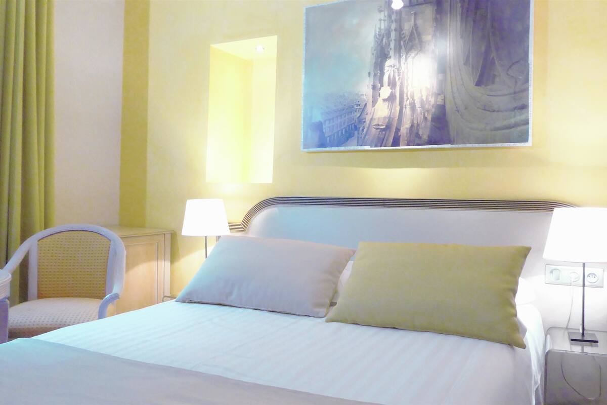 A hotel room with a bed, two bedside lamps, a chair, and a painting on the wall.