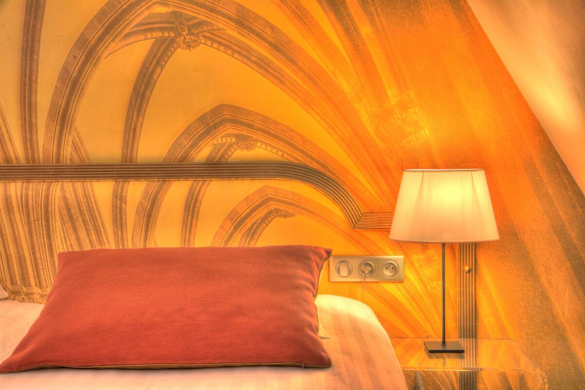 A hotel room with a bed, a red pillow, a nightstand, and a lamp.