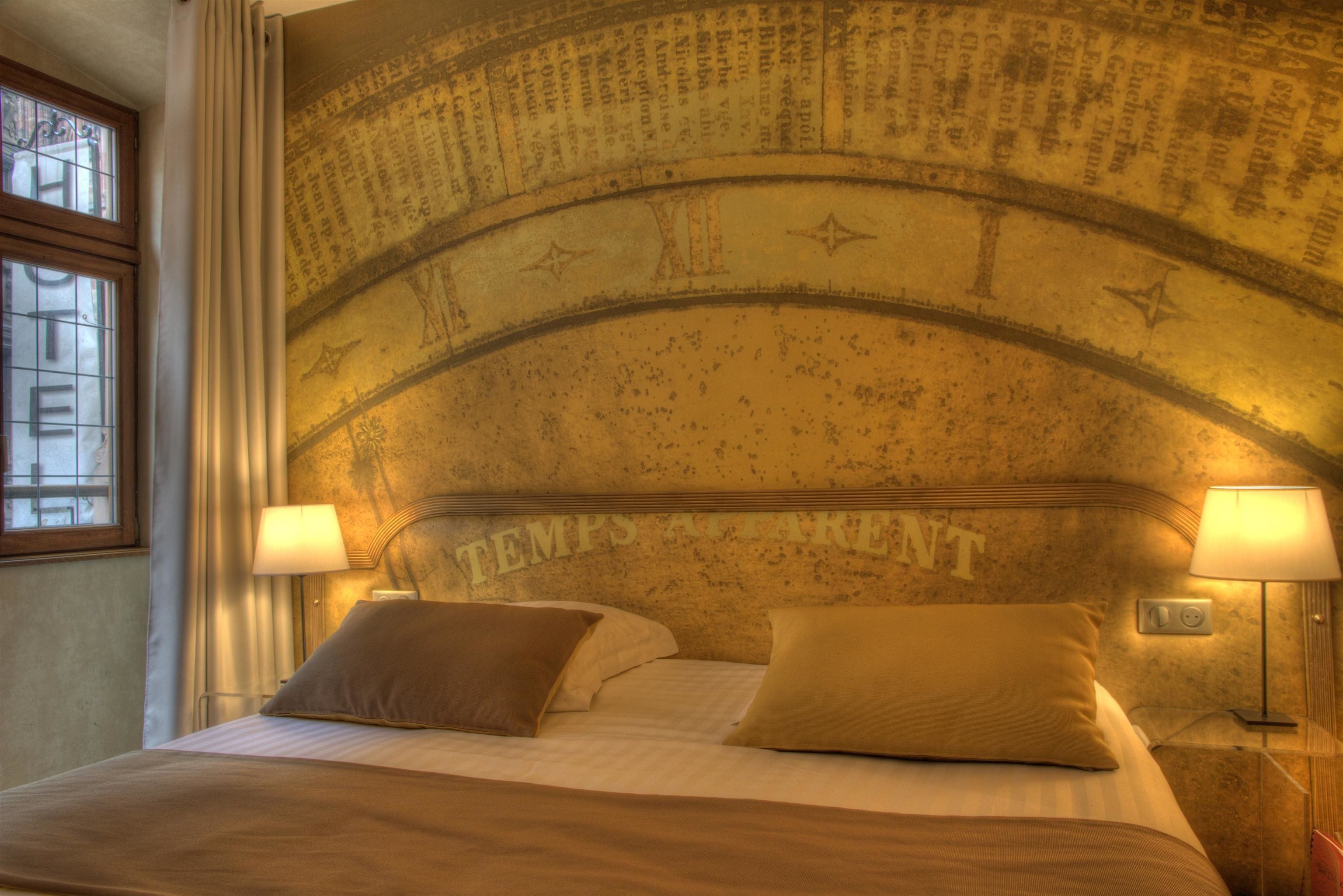 Une chambre d’hôtel avec un grand lit, deux lampes de chevet et un mur orné d’une arche décorative ainsi que des inscriptions indiquant « TEMPS À TARENT ».