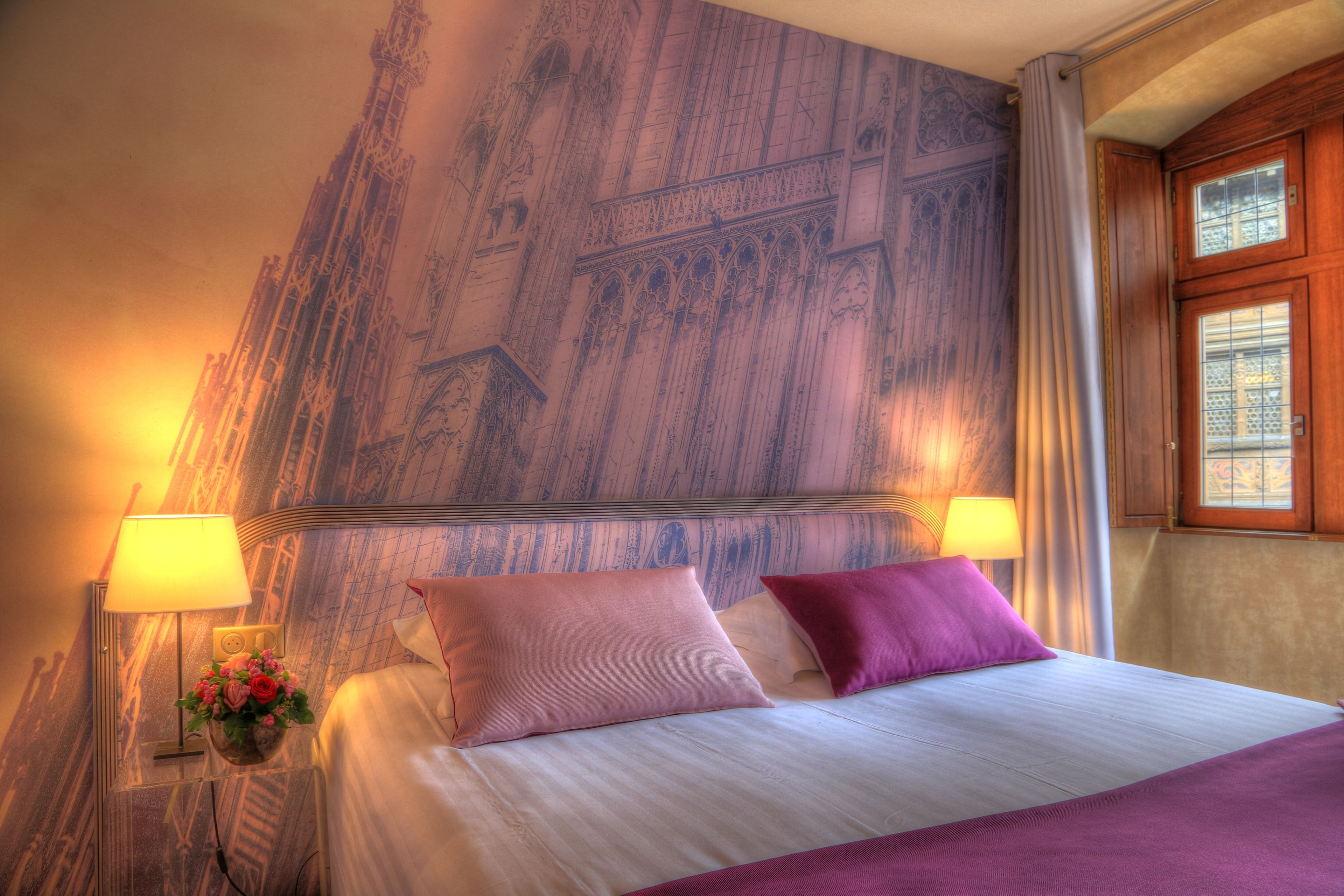 Une chambre d’hôtel avec un grand lit, deux lampes de chevet, une table de chevet ornée d’un vase de fleurs et une fresque murale.