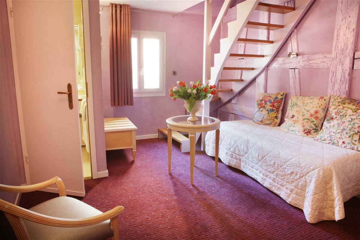 A bedroom with a bed, a chair, a small table with a vase of flowers, a staircase, and a door leading to another room.