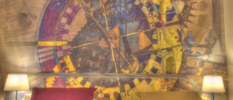 A bed with two pillows, a headboard with a clock, and a wall with a map design.