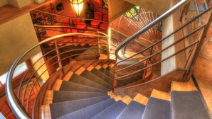 Un escalier en colimaçon avec des rampes en métal et des marches recouvertes de moquette, menant à un niveau inférieur où se trouvent des chaises rouges et une table.