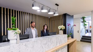 A reception desk with a marble top, two people standing behind it, and a vertical wooden panel wall. - Citadines Wilson Toulouse (Toulouse)