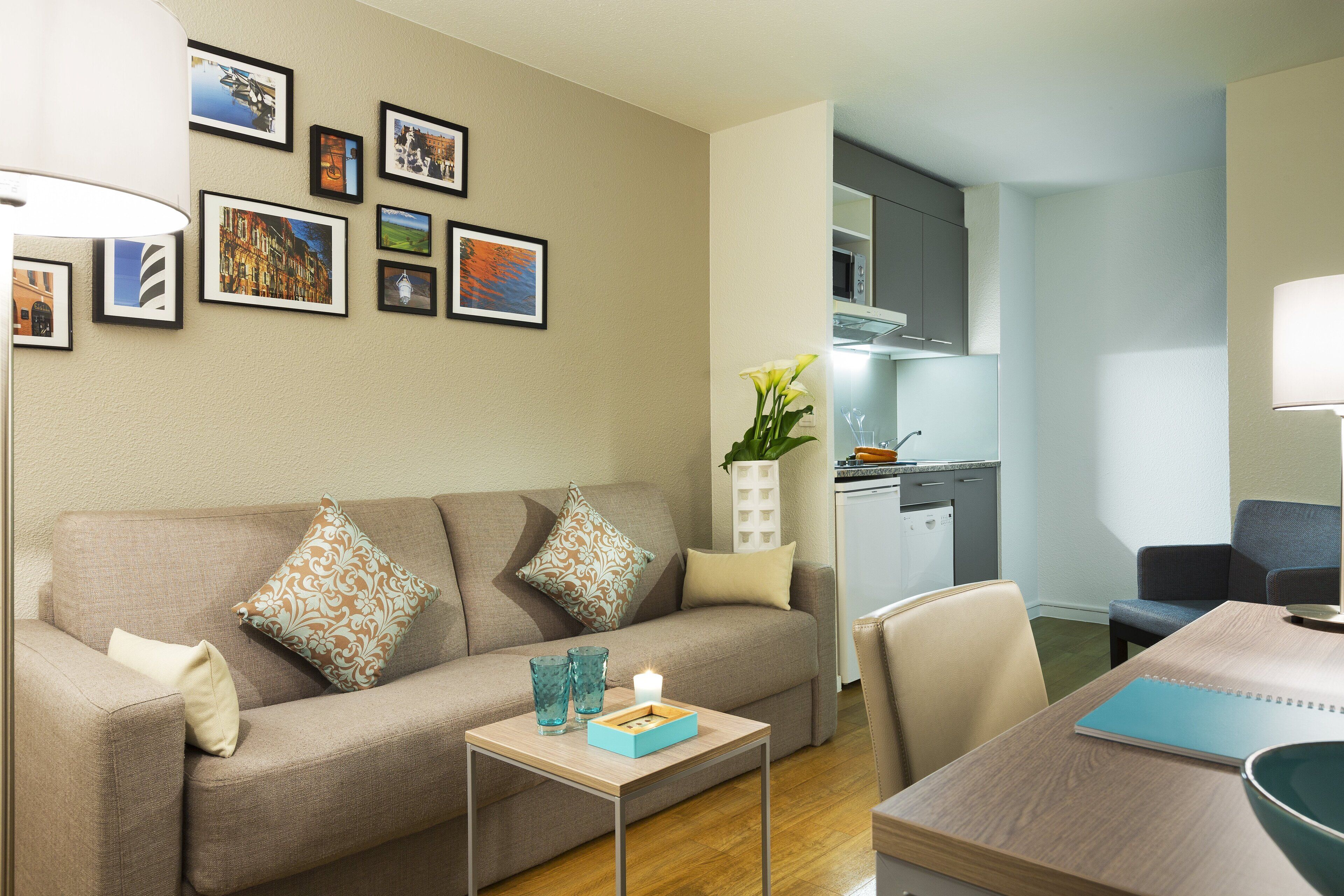 A modern living room with a sofa, coffee table, and a dining area. There are framed pictures on the wall and a kitchen area in the background.