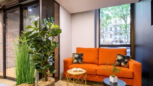 A modern living room with a brick wall, a large window with a city view, an orange sofa with decorative pillows, and a round wooden side table with a book and a potted plant. - Citadines Wilson Toulouse (Toulouse)