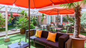 An outdoor seating area with a sofa, chairs, and tables under orange umbrellas, surrounded by potted plants and a greenhouse in the background. - Citadines Wilson Toulouse (Toulouse)