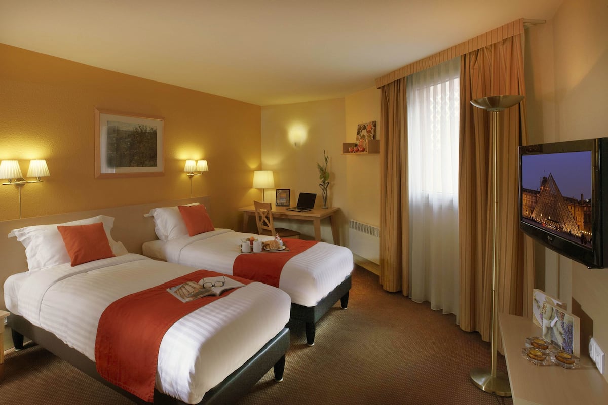 A hotel room with two beds, a desk, a chair, a TV, and a window with curtains.