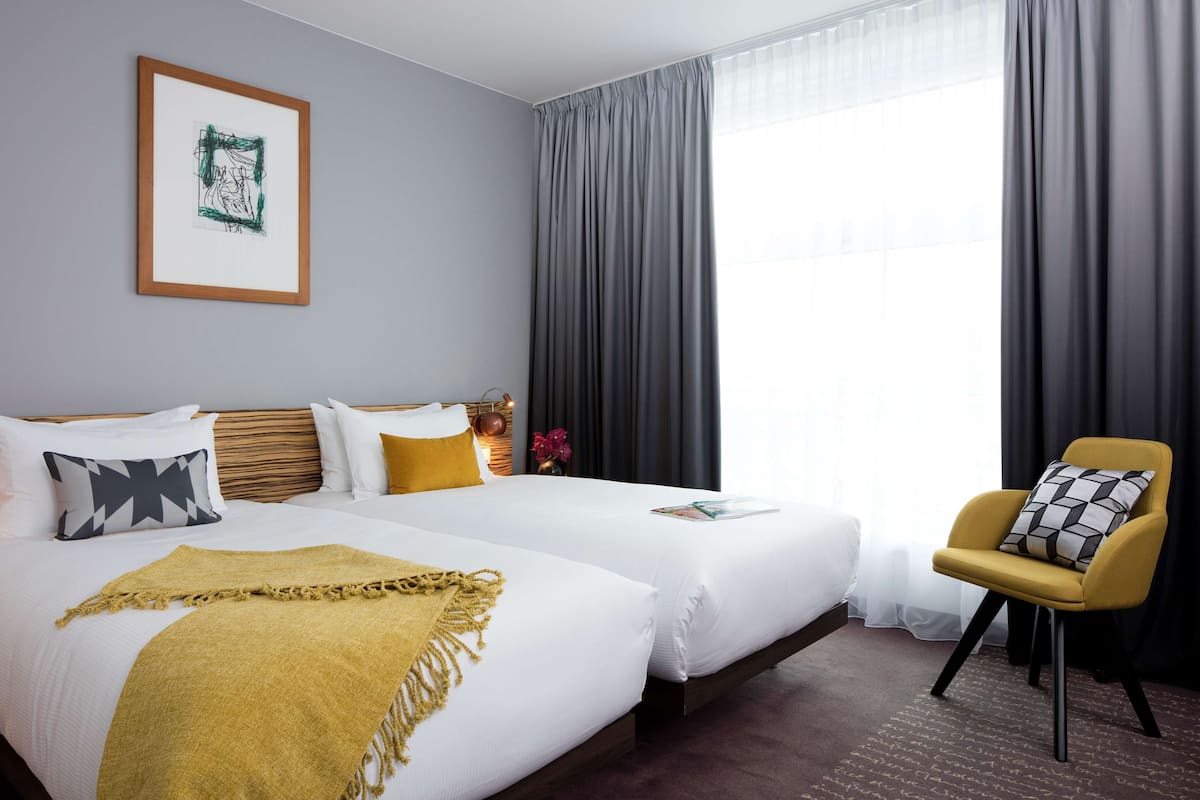 A hotel room with two beds, a framed artwork on the wall, a yellow armchair, and a large window with curtains.