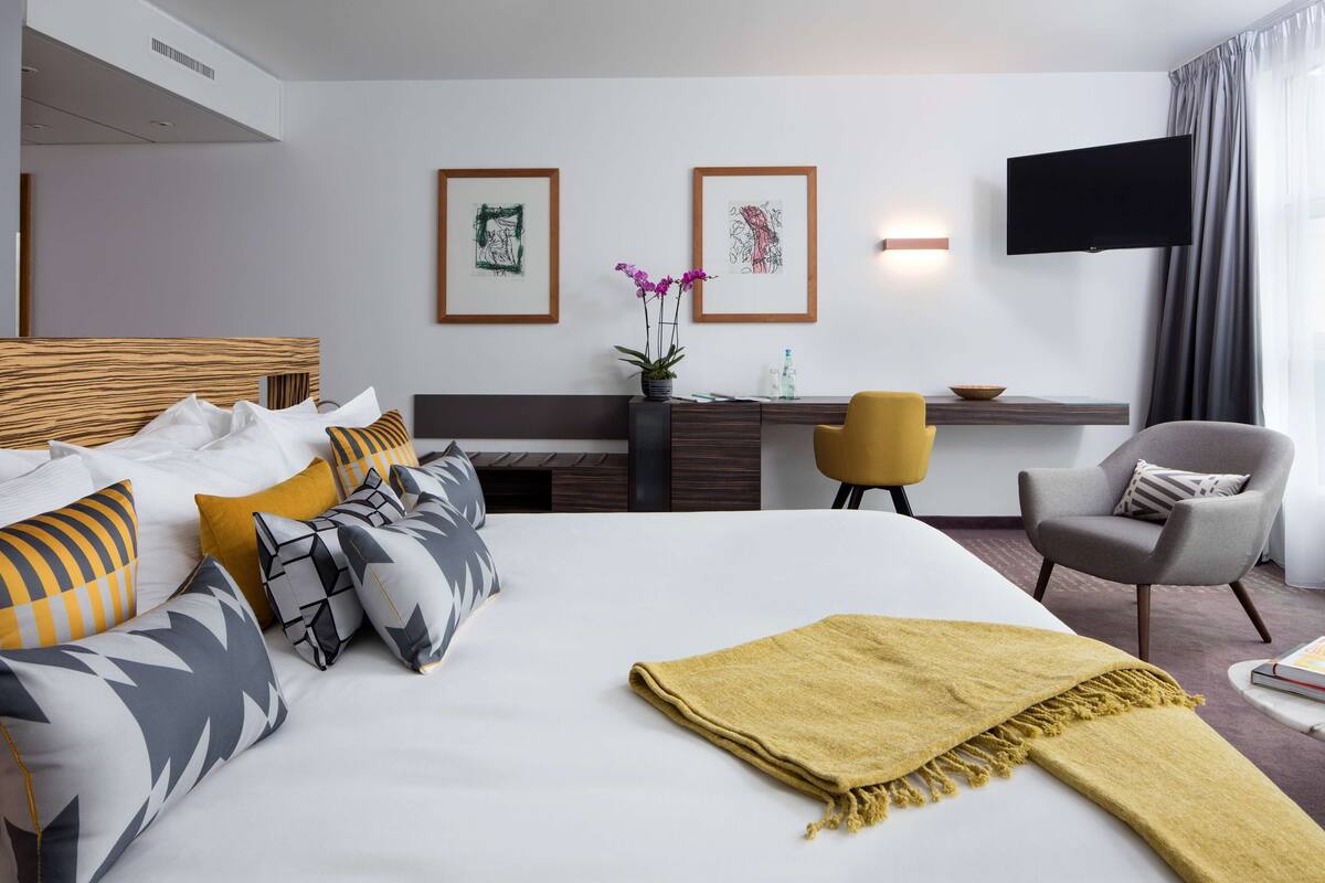 A hotel room with a large bed, a desk, a chair, a television, and artwork on the wall.