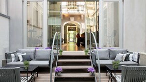 A modern outdoor seating area with grey sofas and chairs, a wooden staircase leading to an interior space, and purple flowers as decoration. - art'otel Berlin Mitte (Berlin)