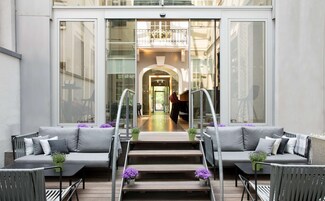 Un'area esterna moderna con divani e poltrone grigi, una scala in legno che conduce a un ambiente interno e fiori viola decorativi.