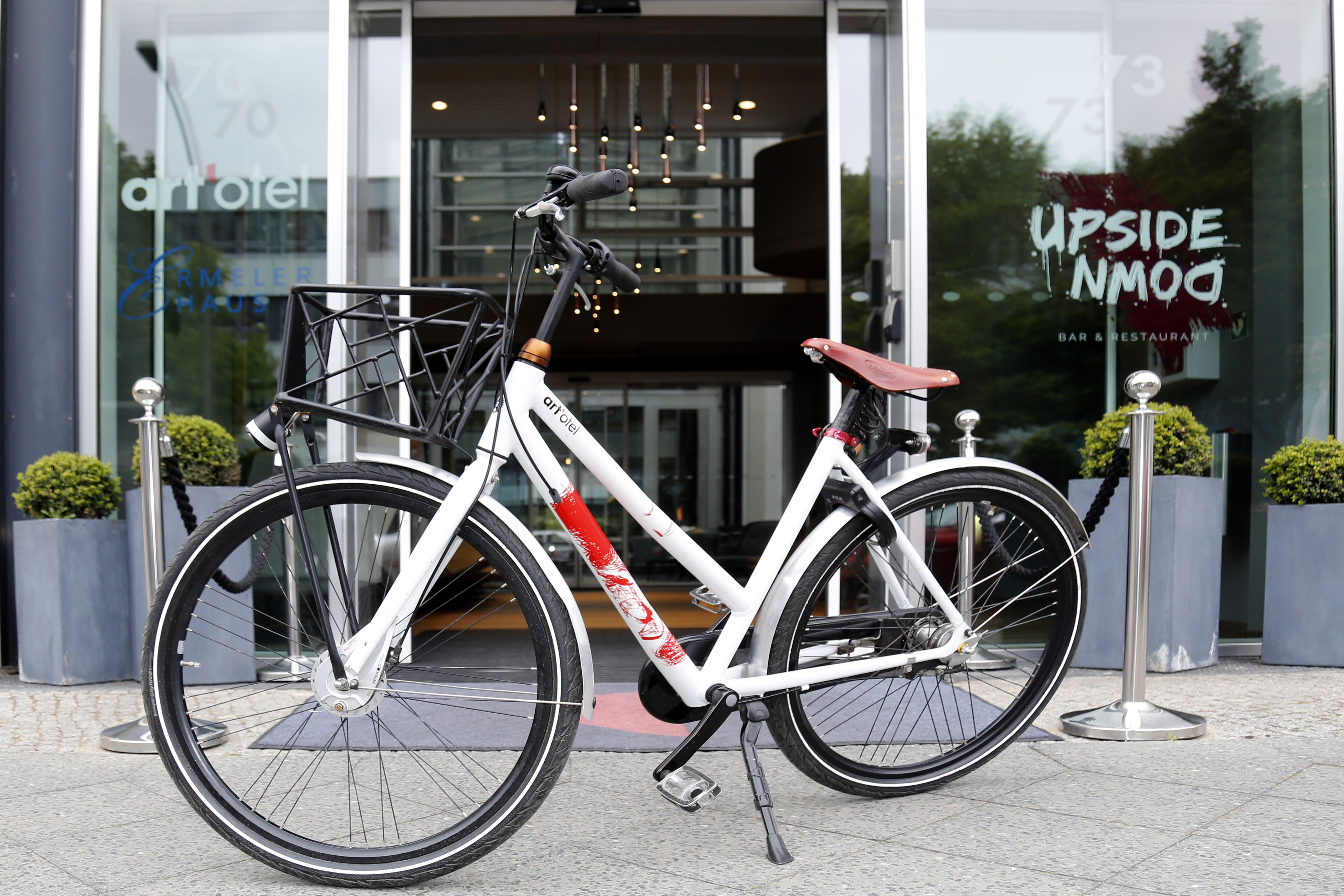 Ein weißes Fahrrad mit Korb und rotem Sattel vor einem Gebäude mit Glastüren.