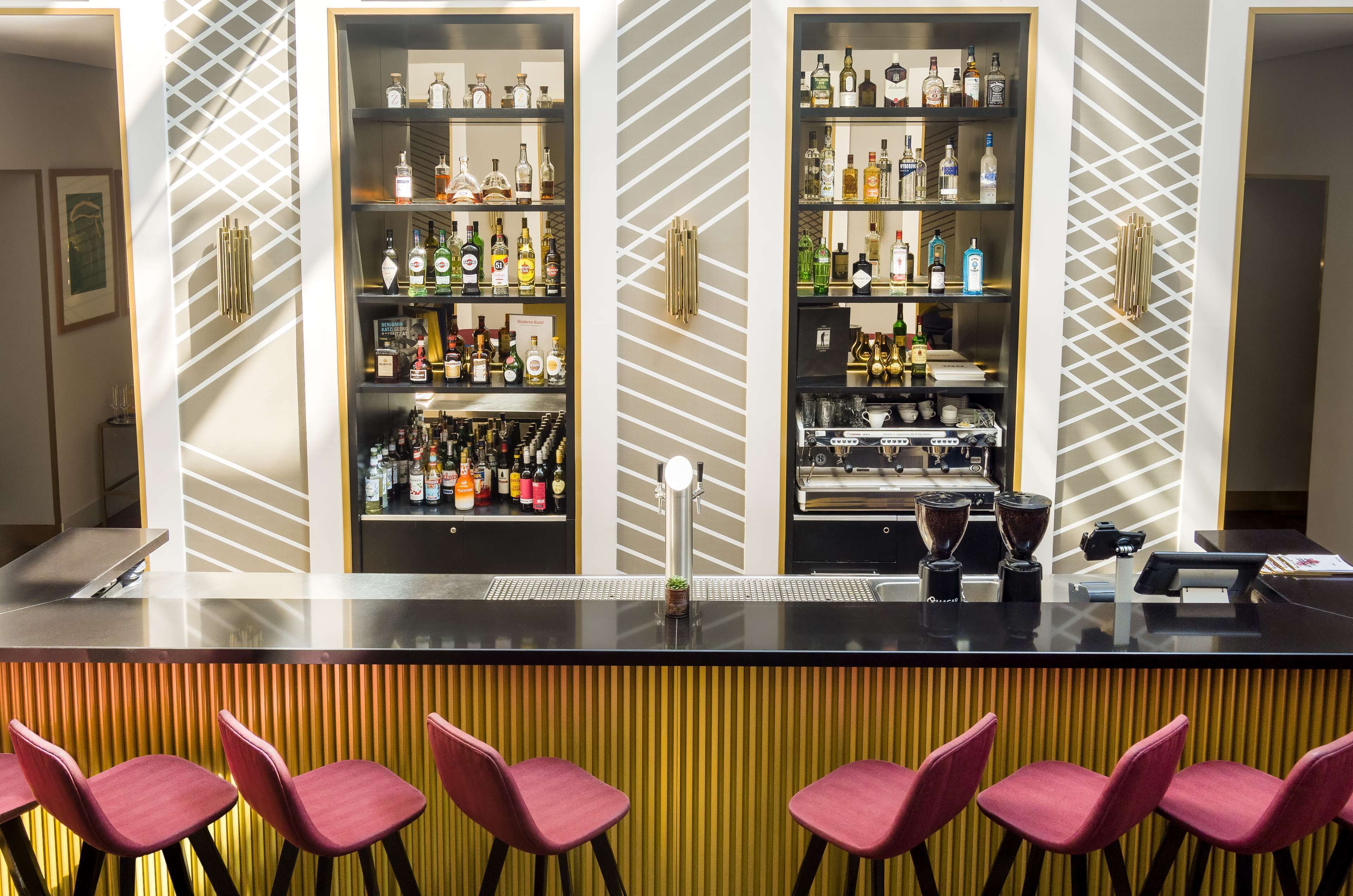 A bar area with a modern design, featuring a black countertop, red bar stools, and shelves stocked with various bottles and glasses.