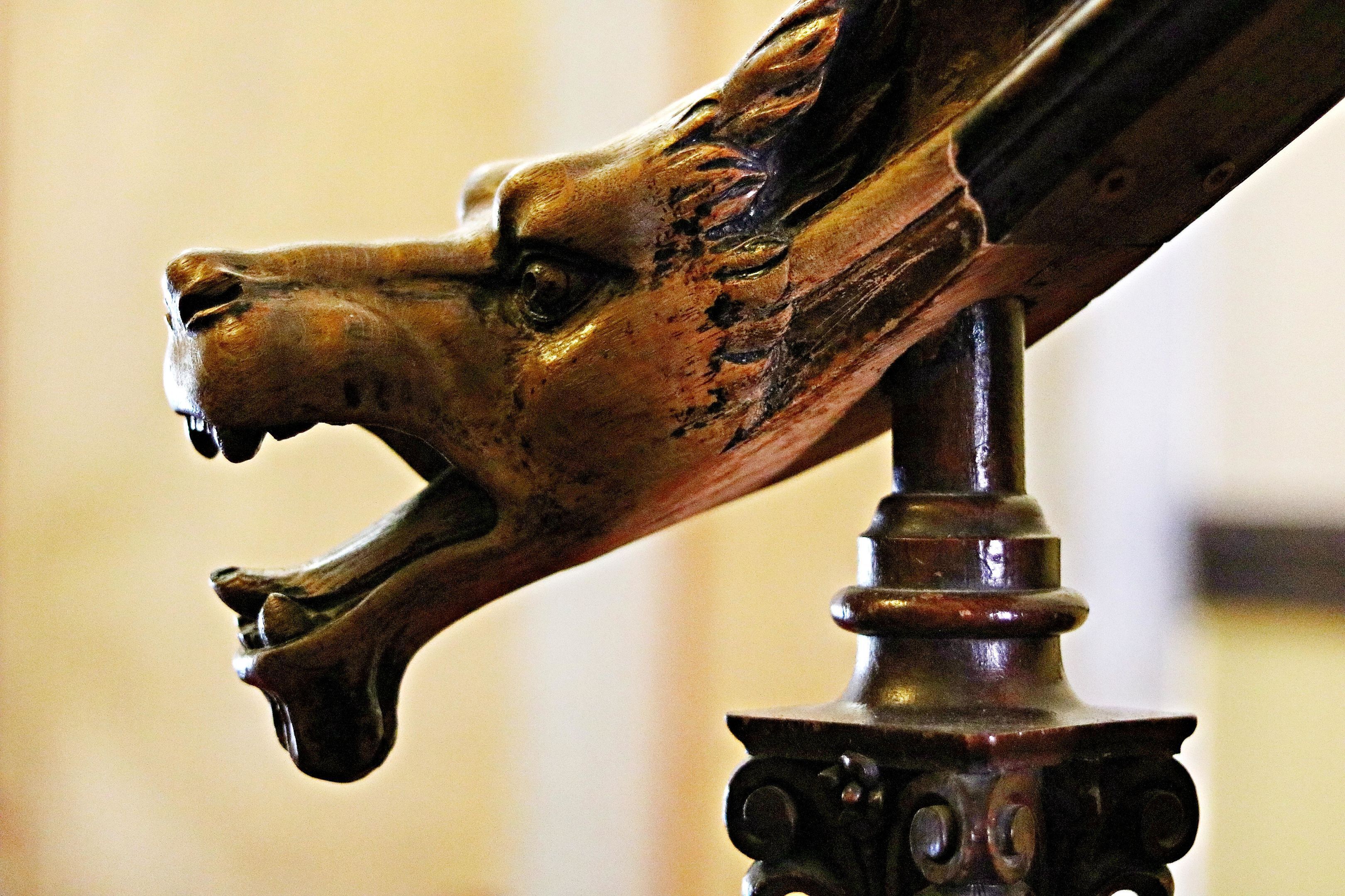 Primer plano de una escultura de madera con cabeza de animal tallada, posiblemente un lobo, sobre un pedestal oscuro y ornamentado.