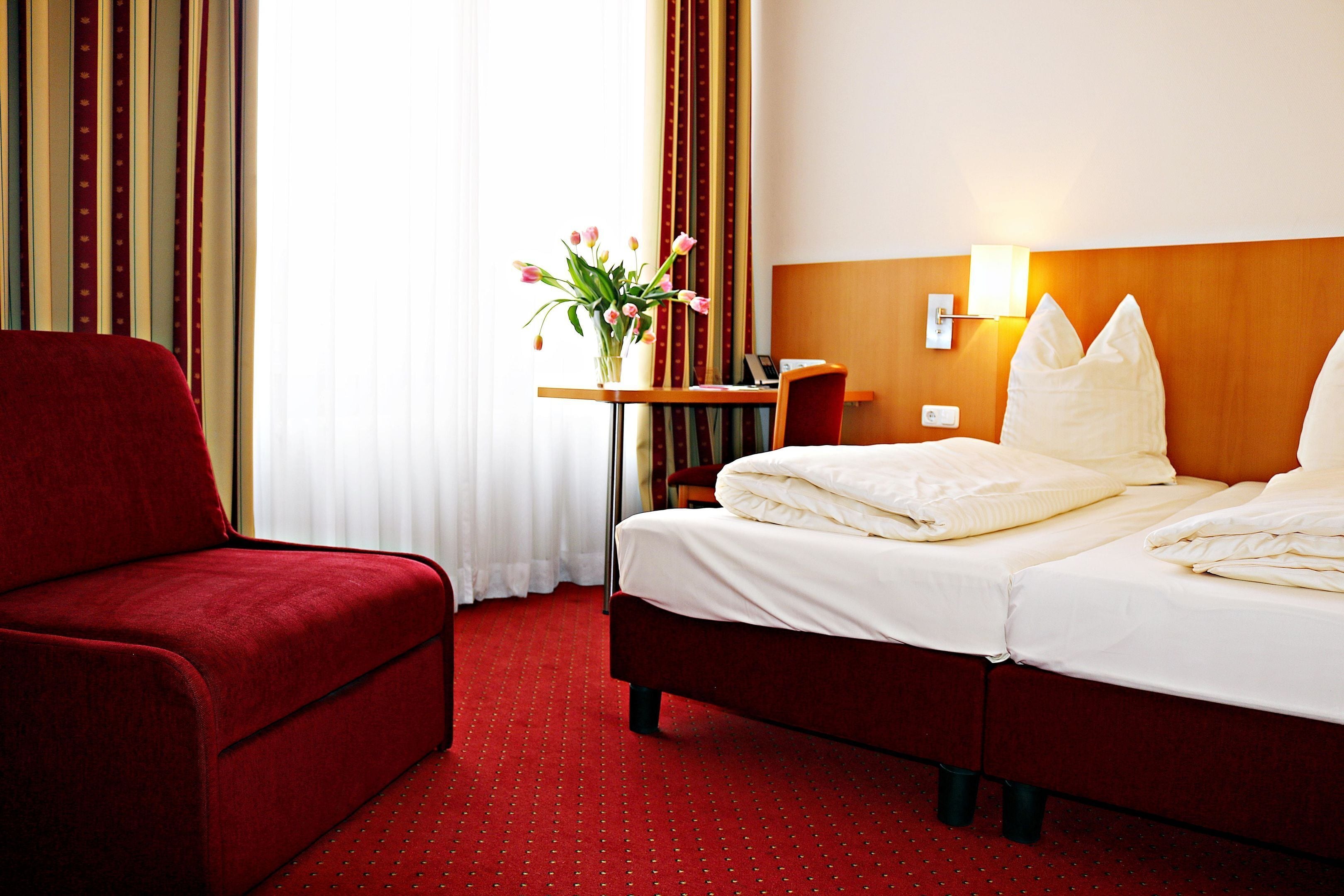 A hotel room with a red sofa, a bed with white linens, a small table with a vase of flowers, and a lamp on the wall.