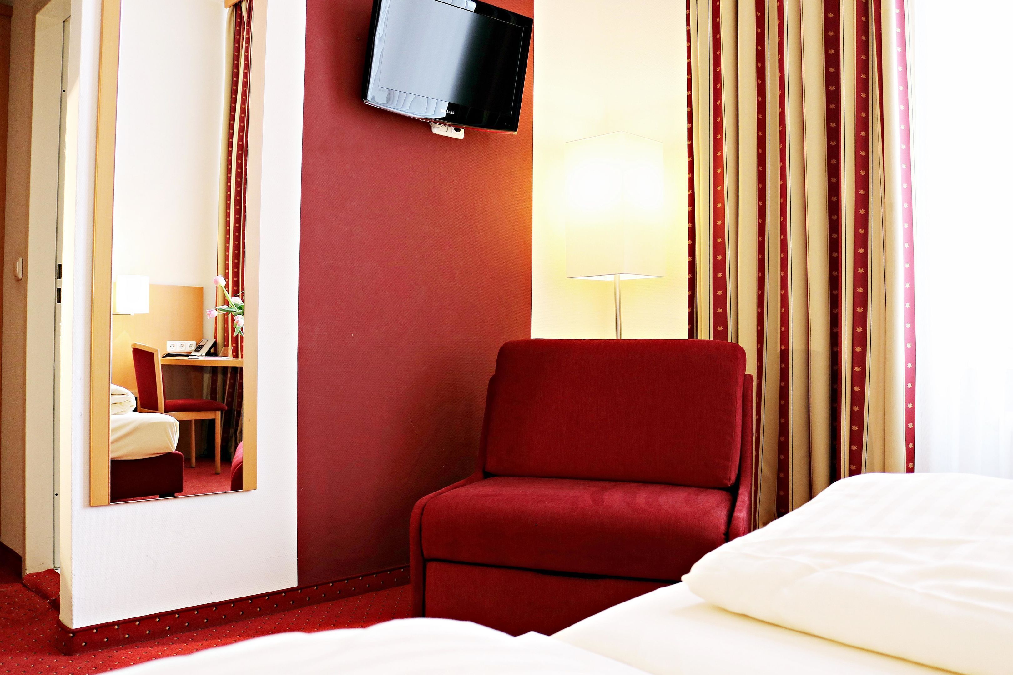 A hotel room with a bed, a red armchair, a television mounted on the wall, and a lamp.